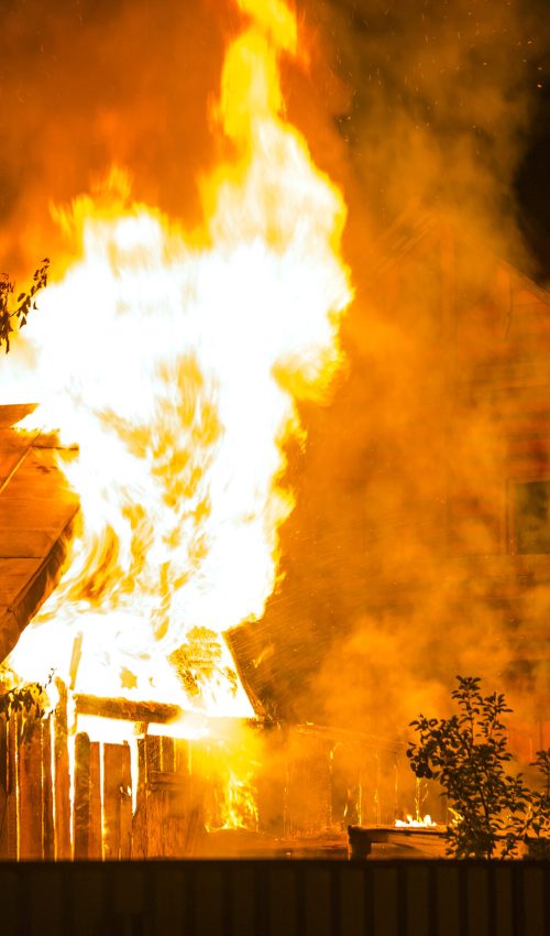 Burning wooden house at night. Bright orange flames and dense smoke from under the tiled roof on dark sky, trees silhouettes and residential neighbor cottage background. Disaster and danger concept.